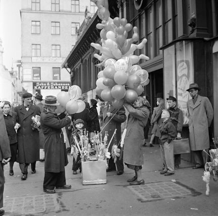 Utanför en äldre saluhallsbyggnad med stora fönsterpartier, står en äldre kvinna och säljer ballonger. Hon har ett stort knippe ballonger i ena handen. Runt omkring är det mycket folk och i bakgrunden syns ett äldre hus med en kaffehandel i bottenvåningen.