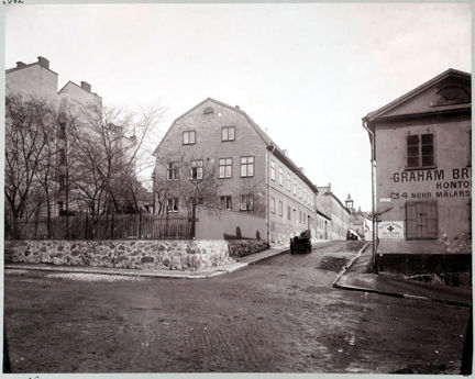Gatubild med korsning. gatsten på vägen, bakom en stenmur och en trädgård ligger ett tvåvåningshus, på andra sidan gatan ser vi gaveln av ett annat äldre hus med reklam på väggen: Graham brothers, kontor.