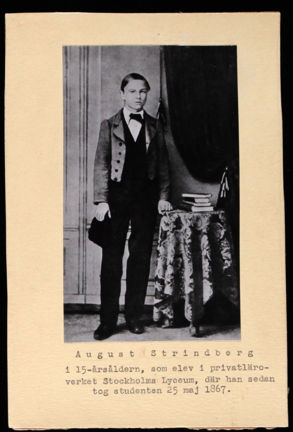 En svartvit bild av August Strindberg som står vid ett runt, mindre bord. På bordet ligger en trave med böcker.