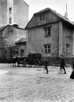 En liten smal byggnad av trä med två våningar och inredd vind står i en gatukorsning. I bakgrunden syns ett högre och relativt nybyggt stenhus. På gatan passerar en häst med vagn framför trähuset. Människor promenerar förbi.