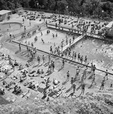 Uppifrån ett högt berg ser vi ett poolområde med två bassänger, en stor och rektangulär, en liten och rund. På solterrasser av trä ligger människor och badar. Det är gott om folk både i och omkring bassängerna.