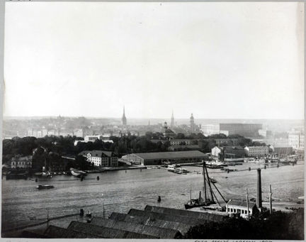 En utsiktsbild mot Skeppsholmen. Här syns ett gammalt badhus, förråd och båthus. I bakgrunden Gamla stan och slottet.  