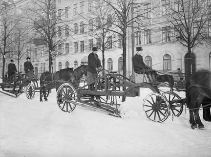 Flera ekipage med hästförspända snöplogar. Kala träd och bebyggelse i bakgrunden.