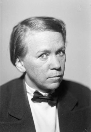 Svart-vitt portätt med ett kvinnoansikte i centrum. Kvinnan har håret i sidbena, tittar in i kameran och bär kavaj och någon slags fluga.