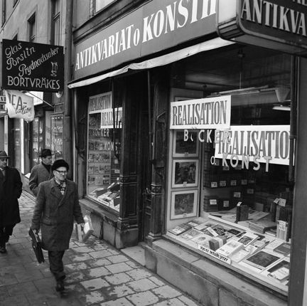 Skyltfönster med anslag om realisation. Tre män passerar på trottoaren.