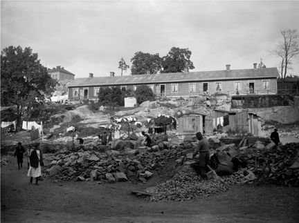I fonden uppe på en kulle syns en huslänga med flera bostäder. Nedanför arbetar människor med olika syssor. 