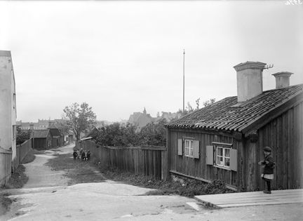 Svart-vit bild med ett lågt trähus och ett barn i förgrunden. Nerför gatan går ett lågt träplank med gräs och träd längs sidorna.