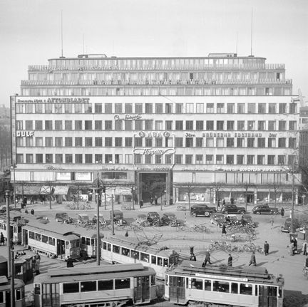 Ett stort hus i totalt 8 våningar, men ljus fasad, många fönster och flera takterrasser, ligger vid ett större torg. På torget möts flera spårvagnar och en mängd bilar och cyklar står parkerade. På husets fasad syns flera ljusreklamskyltar.