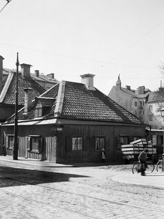 Ett enplans trähus på hörntomt. Spårvagnsspår i gatubeläggningen, några cyklar står parkerade vid trottoaren. En person skyndar förbi.
