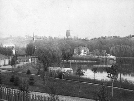 I lummig miljö på andra sidan om ett större vattendrag ligger en äldre vit stenbyggnad. På berget ovanför står en väderkvarn.