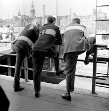 Tre män lutade mot ett räcke vid Slussen. Gamla Stan till vänster i bakgrunden, till höger Saltsjörampen