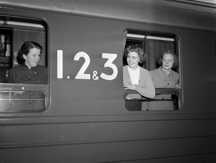 Tre kvinnor tittar ut genom öppna fönster på en tågvagn märkt "1. 2 & 3".