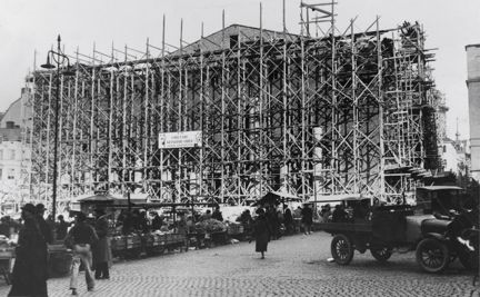 Hus under uppbyggnad, byggnadsställningen i trä framför. I förgrunden Hötorget med några människor och en bild.