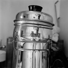 Självporträtt speglat i rostfri kanna. Fotografen befinner sig i en köksinteriör, som också speglas i kannan