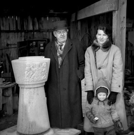 Svartvitt fotografi, porträtt av tre personer med en hög dopfunt i sten.
