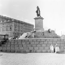 Skeppsbron, Gustav III:s staty. Sittande människor på trappstegen. I bakgrunden Stockholms slott