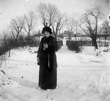 Vinter. En kvinna i hatt och lång kjol och kappa tittar in i kameran. i bakgrunden träd och ett hus.