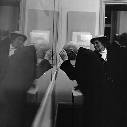 En äldre man i mitten av bilden lutar sig fram emot en musei-monter. Han speglas i monter-glaset till vänster i bild. I bakgrunden syns även en kvinna i hatt.