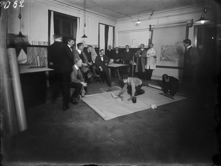 Svart-vit bild infrån ett rum med vita väggar och trägolv. På golvet ligger ett stort papper som två små pojkar sitter på. Runtom pappret står en grupp om totalt tolv män. Männen bär rock eller kostym med slips. På väggen till höger hänger en karta.