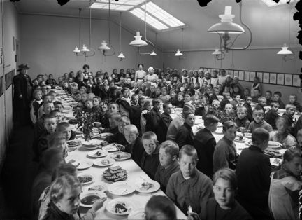 många skolbarn sitter vid långbord med tallrikar med mat framför sig.