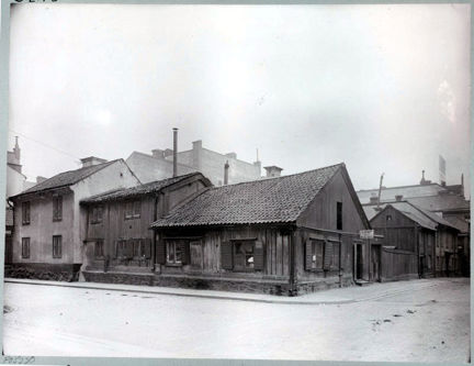I ett gathörn ligger flera små, låga träkåkar runt en innergård. I bakgrunden anas höga, nybyggda stenhus.