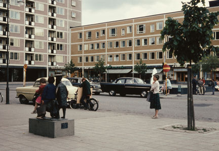 Vid ett litet torg syns ett höghus och ett lägre hus. Med i bild finns även träd, en fontän, ett par bildar, en kvinna som ser ut att stå i egna tankar och några barn som klättrat upp och satt sig på en skulptur.