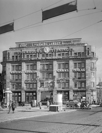 Centralpalatset vid Tegelbacken. Fasaden täckt av reklamskyltar i neon. Människor flanerar, bilar på gatorna.