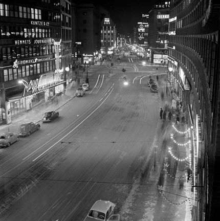 Bilden är tagen från en höjd. På fasaderna sitter många neonskyltar, bland annat över biografen Royal och varuhuset Pub. Ljusa streck längs gatan indikerar en lång slutartid, som gjort att bilars lysen lämnat spår, utan att bilarna syns.