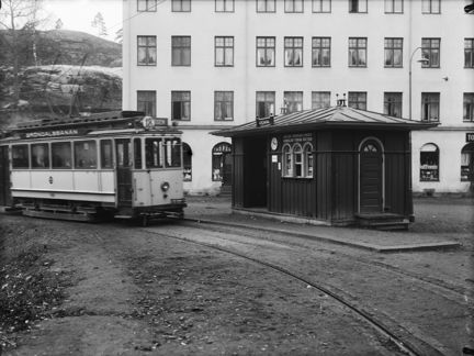 Spårvagn framför stort bostadshus och med en liten träbyggnad intill.