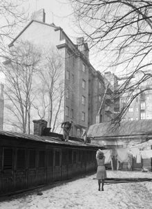 Kammakargatan 70, gården. En kvinna hänger tvätt och två män arbetar på taket till längan med utedass. Huset i fonden ligger vid Tegnérgatan 57