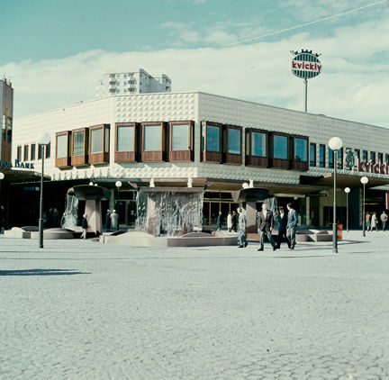 Ett stenbelagt torg med flera fontänskulpturer. I bakgrunden affärsbyggnad med vit fasad och burspråk av brunt trä. Flera män går över torget och en liten pojke står intill fontänen.