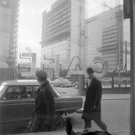 Två personer, en bil och Hötorgshusen under uppbyggnad. Vy genom caféfönster.