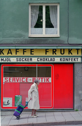 Kvinnan drar en dramaten. På butikens skyltar står: "Kaffe. Frukt. Mjöl. Socker. Choklad. Konfekt. Service-butik.")
