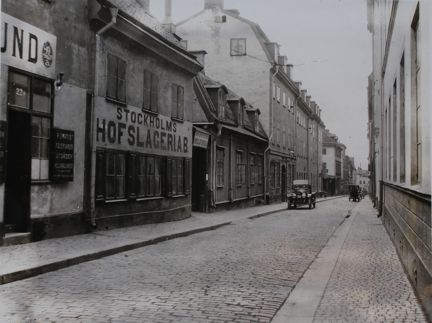 Äldre hus i ett par våningar kantar en gata belagd med gatusten. På fasaderna är butiksnamn målade eller omnämnda på skyltar. I fonden två äldre bilar.