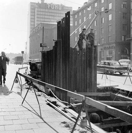 Två män står på järnbalkar i en grop i gatan. Runt gropen finns en avspärrning. I bakgrunden syns Konserthuset och en av Hötorgsskraporna. På skrapan finns en stor reklamskylt för Ford. 