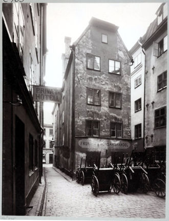 Ett högt och smalt hus  med grund från medeltiden ligger mellan två smala gränder och ett litet torg. På torget är kärror uppställda. Från fasaden hänger en skylt ut som visar att det ligger ett hotell i fastigheten.