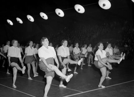 I en gymnastiksal, under en rad med runda lampor, syns kvinnor i gymnastikkl&#xE4;der utf&#xF6;ra gymnastikr&#xF6;relser. Kvinnorna &#xE4;r kl&#xE4;dda i vita blusar och sportiga shorts.