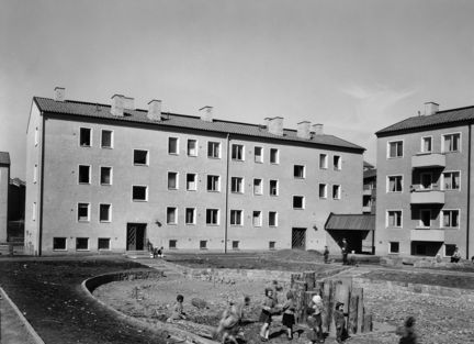 Två flerfamiljshus i tre våningar, delvis förbundna av ett litet skärmtak. Mellan husen syns en ännu ej färdig gård med en liten lekplats och lekande barn. 