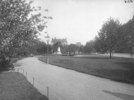En park med grusgångar och gräsmattor, träd och planteringar. En bit bort står en skulptur av en man på en sockel och bakom honom skymtar gaveln av en stationsbyggnad.