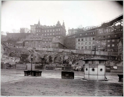 Större och mindre stenhus klättrar på ett högt berg. En hög gångbro till höger i bild. I förgrunden en mindre kur med snickarglädje.
