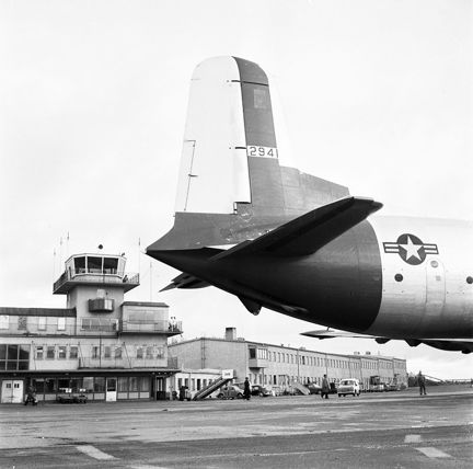 Vi ser stjärten på ett större passagerarflygplan som står parkerat framför flygterminalen och flygledartornet, vilket syns i bakgrunden.