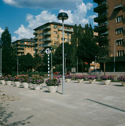 Rader av blomsterurnor, tunnelbaneskylt, funkishus i bakgrunden.