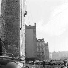 Rivningstomt med parkerade bilar i kv. Uppvaktaren. Arbetare upphissad med linor framför brandmur med plåttäckta rökgångar