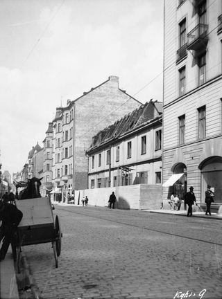 En stenbelagd gata. till vänster syns en häst med vagn, till höger äldre träbebygglse och högre stenhus. 