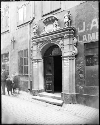 En port med dubbeld&#xF6;rrar, omsluten av en stenportal med skulptural utsmyckning. Utanf&#xF6;r p&#xE5; gatan st&#xE5;r flera barn och tittar &#xE5;t fotografens h&#xE5;ll. Den ena d&#xF6;rren st&#xE5;r &#xF6;ppen.