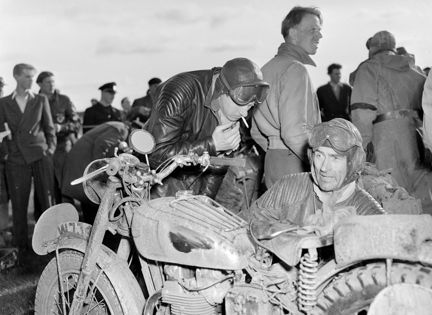 fullt med människor på en racerbana. några män meckar med en motorcykel, den ena har en cigarett i munnen.