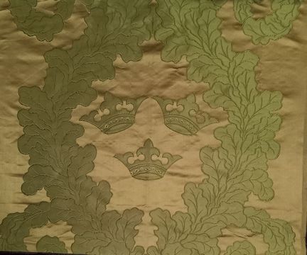 Bilden visar ett grönt sidentyg. På tyget finns tre kronor som är omslutan av en eklövskrans. Tyget i botten är ljusare än kransen och kronorna.