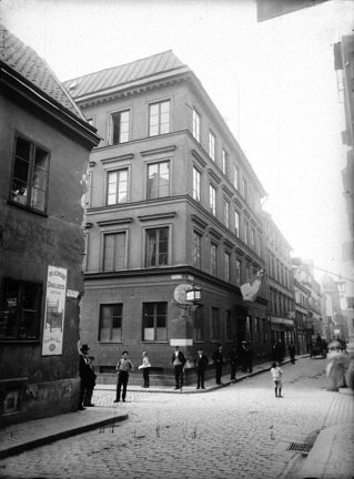 Vid en gatukorsning står en del människor och blickar mot fotografen. De står framför två hus, den bortre större än det närmast i bild. Gatan är belagd med gatsten. På det större huset hänger en flagga ut från fasaden. 