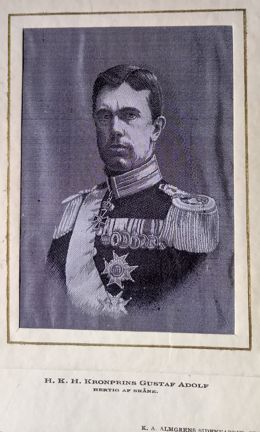 En vävd bild av kronprinsen Gustaf Adolf. Prinsen ser sträng ut. Han har sidbena. Han har på sig uniform med 7 medaljer på bröstet och en dekoration runt halsen. Han bär också två stora ordersstjärnor (troligtvis serafimerorden) och ett ordensband (serafimer). Prinsen har epåletter på axlarna med många fransar. 