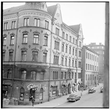 Ett fyra våningar högt stenhus med rundat hörn, krönt av en tornhuva. I bottenvåningen syns butikslokaler. På gatan utanför står bildar parkerad. En man går strax in genom butiksdörren på hörnet.